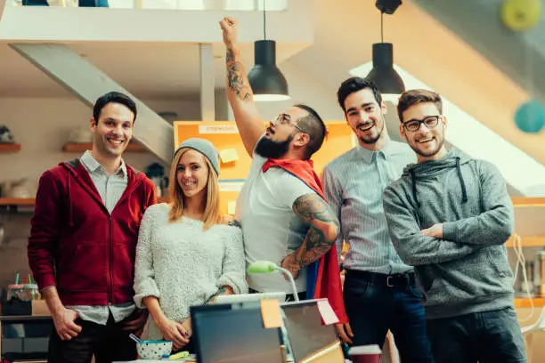 Five young people running their developers startup company. Working in the house like business incubator. Standing and posing for group portrait in their office. Looking at camera and smiling. One of the founders wearing super hero cape and holding his arm raised. Shot with Canon EOS 5Ds.