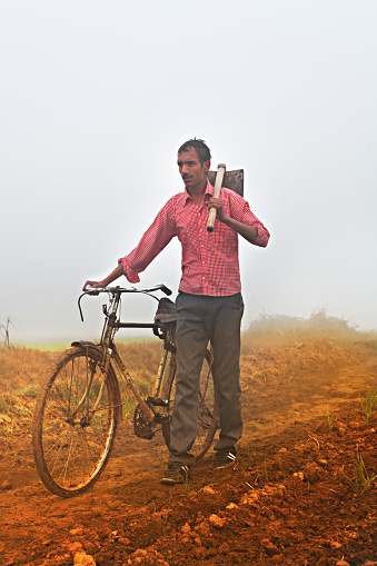 Young Farmer going through the field with his bicycle during winter season wearing red color shirt, he is carrying hoe on his shoulder & looking in front of himself vertical portrait outdoor.