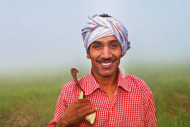 Confident Cheerful Young Indian Farmer Standing in the field holding sickle on his shoulder wears red color shirt & turban on his head, in a foggy day during winter season. He is laughing while standing in the field outdoor portrait close up.