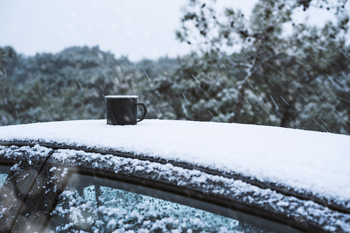 Cup of tea or coffee on the roof of a car, snowy day. Car is covered with snow in a forest.