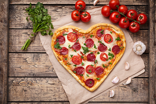 Heart shaped pizza with pepperoni, tomatoes, mozzarella, garlic and parsley composition on vintage wooden table background. Concept of romantic love for Valentines Day. Rustic style. Top view, flat lay.
