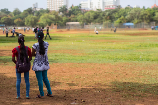 Mumbai, India - June 15, 2014: Two young Indian girls watching a game of cricket in a sports park in Mumbai, India, in the afternoon