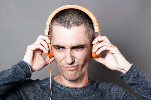 painful sound concept - unhappy 30s man frowning in listening to noise or music on orange headphones,dark texture effects,gray background studio
