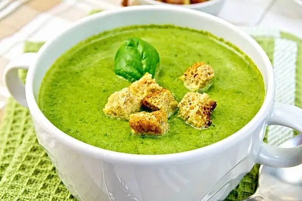 Green soup puree in a white bowl with croutons and spinach leaves on a napkin on the background fabric