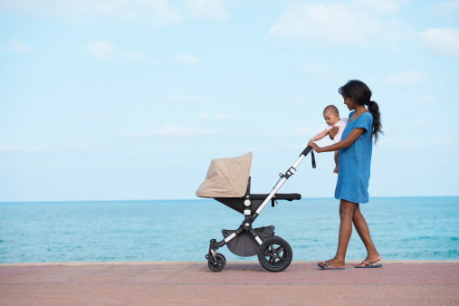 Young mother carrying 6 months old baby and pushing stroller, walking along seacoast.