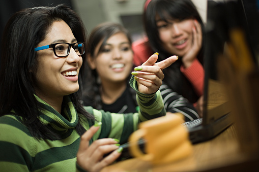 Indoor image of a group of happy late teen girls from different ethnicity using laptop together. Selective focus is on the girl sitting in foreground wearing eyeglasses while pointing at laptop screen. Group of people, waist up, tilt, horizontal composition with selective focus.