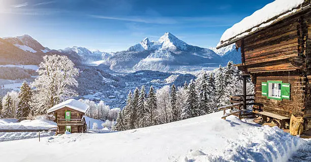 Panoramic view of beautiful winter wonderland mountain scenery in the Alps with traditional mountain chalets on a cold sunny day with blue sky and clouds.