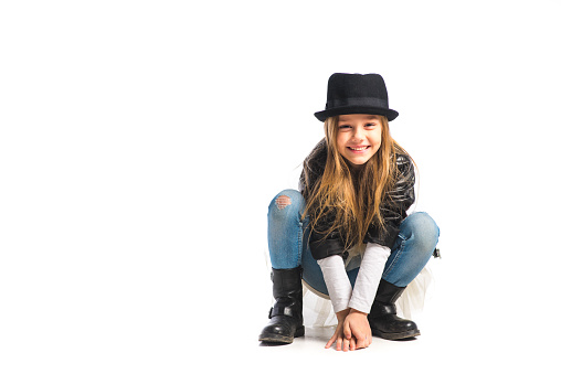 Cute little girl crouching and wearing black hat and black leather jacket. Smiling and looking at camera. Isolated on white.