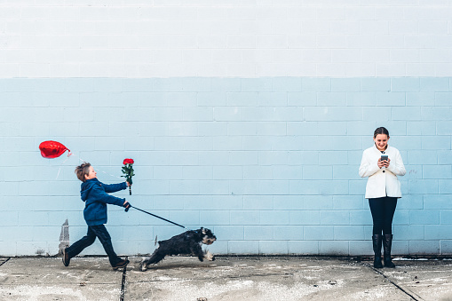 We all need a little help finding love, no matter what tools we use. Little boy with flowers and heart shaped balloon getting pulled by his dog and girl standing using her smartphone looking for something. 