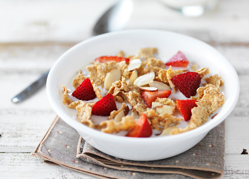 close up of a bowl of cereal and milk with almons and strawvberries on rustic wooden table