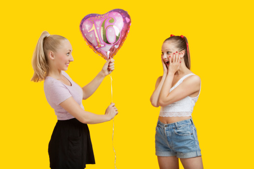 Happy young woman giving birthday balloon to amazed woman standing over yellow background