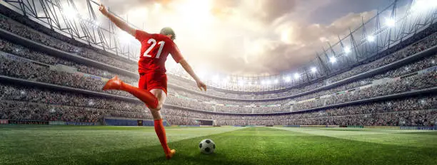 A male soccer player makes a dramatic play by kicking a ball with his feet. The stadium is blurred behind him. It's sunny weather.