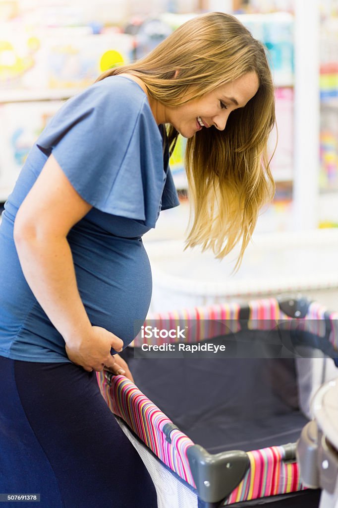 Smiling pregnant woman shopping for crib in baby shop A pregnant woman looks down, smiling, at a camping cot or crib in a baby store. Crib Stock Photo
