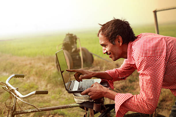 Farmer using laptop in the field Cheerful young farmer of Indian ethnicity standing in the field with his bicycle wearing red checked pattern shirt, and using laptop near green wheat field. He is pointing to the laptop screen and looking happy.  developing countries photos stock pictures, royalty-free photos & images