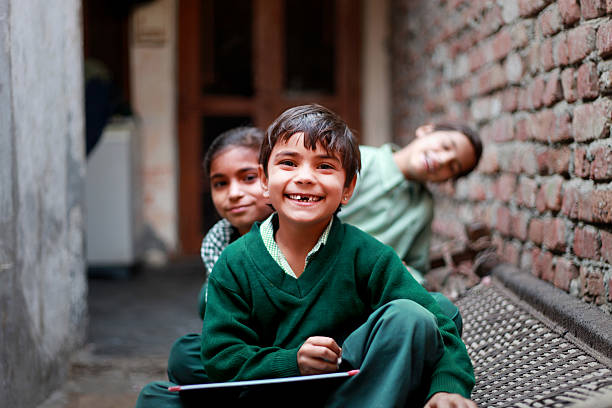 Cheerful School Students of Indian Ethnicity sitting in a row on the Charpai wearing school dress & cheering together. One of them Holding slate & writing with the chalk, they all are laughing together and looking to the camera while sitting at home.
