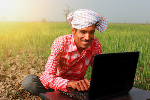 Confident Cheerful Farmer Sitting near Green wheat field & using laptop during winter season in an sunny foggy day, he is wearing red color checked pattern shirt and turban which is traditional dress for men in North India. He is looking cheerful while using laptop in the field, there is sunrise behind him in the field outdoor portrait.