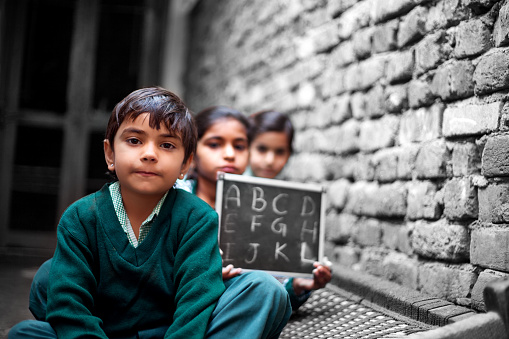 Little School Girls sitting at home near brick wall on Charpai (domestic bed) holding chalkboard wearing school dress and looking to the camera portrait close up.