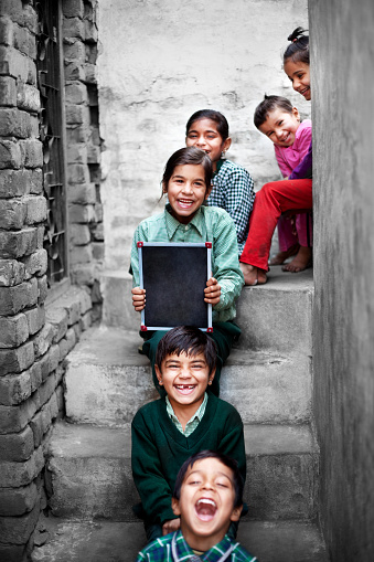 Little School Girls & Boys sitting in the Staircase at home near raw brick wall holding chalkboard (slate) wearing school dress and looking to the camera portrait close up. They all are laughing while sitting in the stairs.