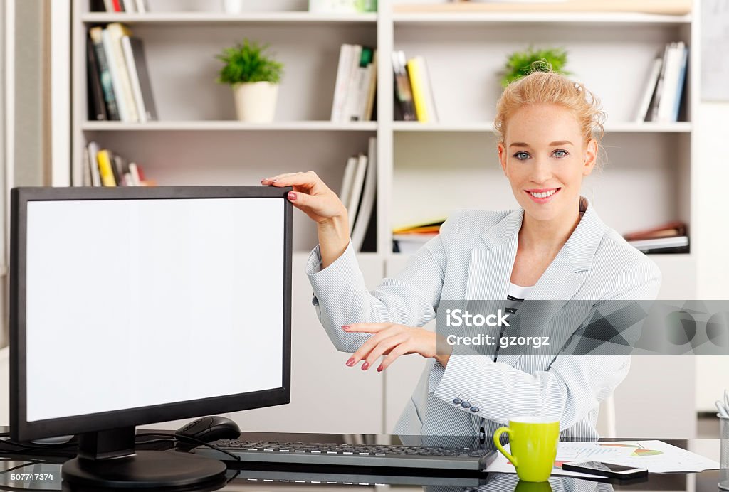 Businesswoman portrait Portrait of young assistant sitting at office in front of isolated white screen computer. Businesswoman showing something with her finger on monitor while looking at camera. 25-29 Years Stock Photo