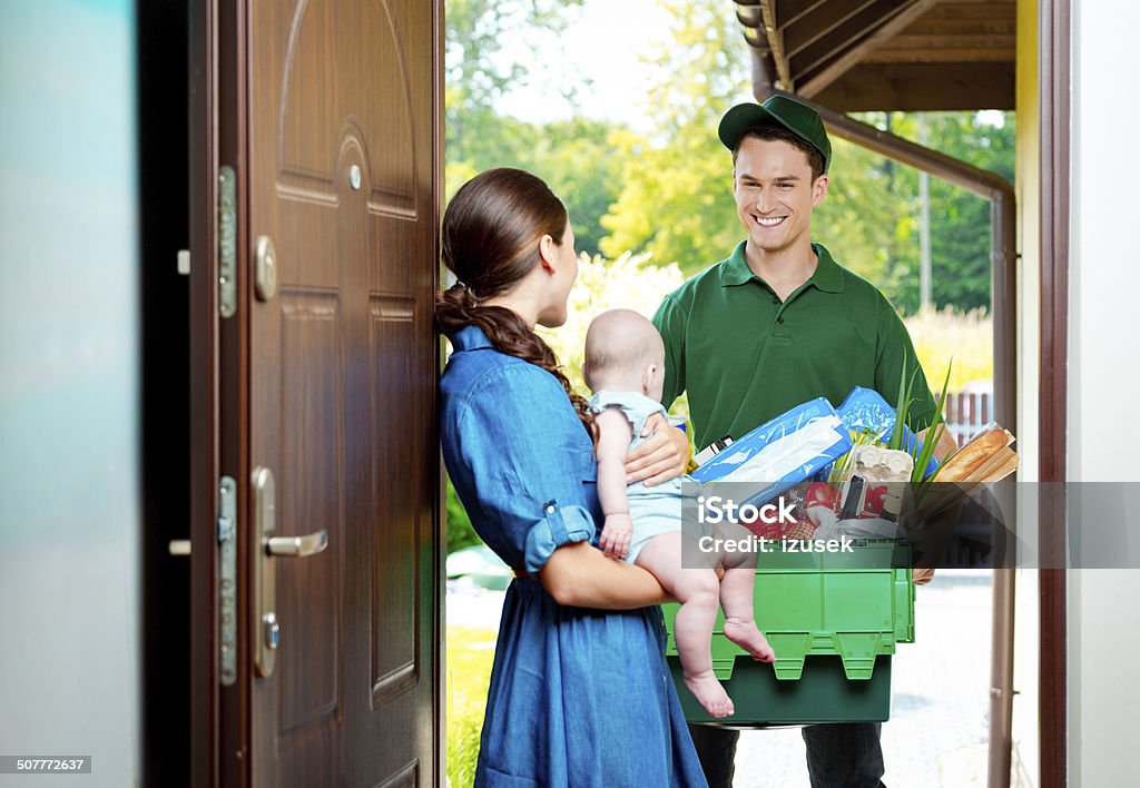 Delivery man with groceries Delivery man standing at the door of the house and carrying box with groceries, talking with woman holding baby. Delivering Stock Photo