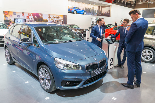 Brussels, Belgium - Januari 12, 2016: Blue BMW 2 Series Active Tourer compact crossover mpv car. The 2 Series Active Tourer is the first Front-engine, front-wheel-drive BMW. Sales men in business suits are standing next to the car. The car is on display during the 2016 Brussels Motor Show. The car is displayed on a motor show stand, with lights reflecting off of the body. There are people looking around and other cars on display in the background.
