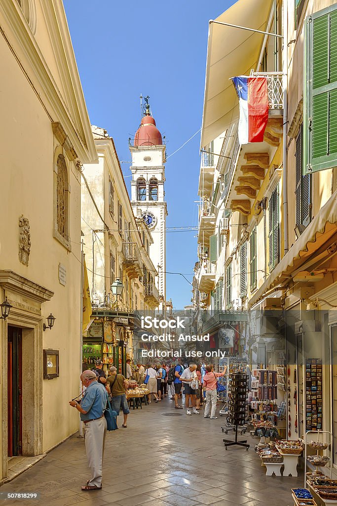 Tourists walking and shopping on narrow streets Kerkyra, Corfu. Greece - September 24, 2013: Tourists walking and shopping on narrow streets with bell tower of Saint Spiridon church in background. It is single-nave basilica and its bell tower is highest in Ionian Islands and most famous church in Corfu Ancient Stock Photo