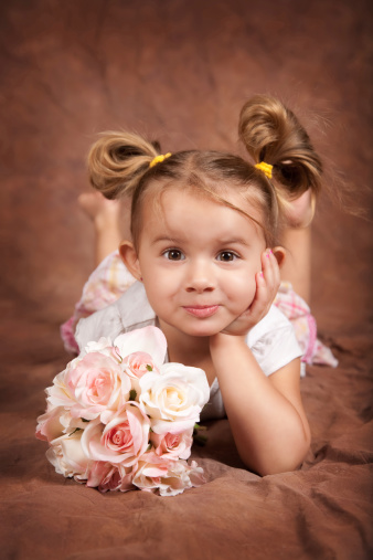 Portrait of 3 year old little girl with brown eyes and sandy blonde hair posing on a brown background.  Holding a boquet of flowers. Child has a silly grin.