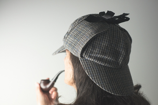 A shallow depth of field portrait shot from over the shoulder of a woman wearing a Sherlock Holmes deerstalker hat and holding a smoking pipe. 