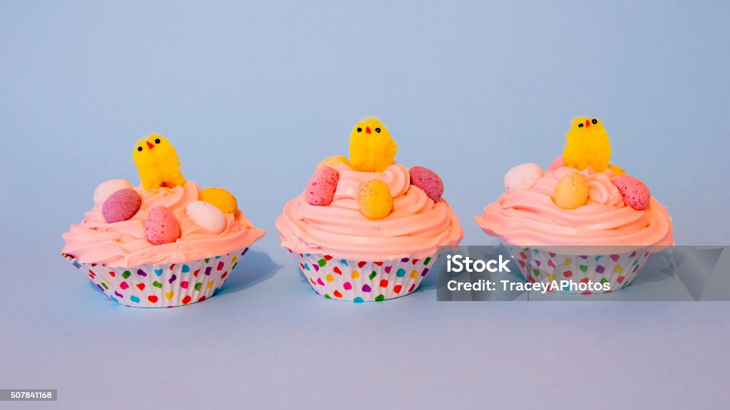 Three Easter Cupcakes with Yellow Chicks and Easter Eggs Three cute Easter cupcakes, with pink buttercream icing, candy Easter eggs, and yellow chicks. Blue background, cakes in multicolored cases.  Animal Stock Photo