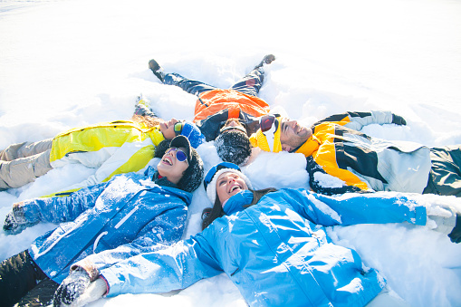 Group of five happy friends lying in a circle on the snow. Some od them wearing goggles. Concept for hapiness and fun.