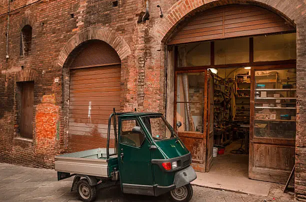 An dark green Ape car parked in front of a repair shop in an old street of Siena, in the Italian Tuscany.