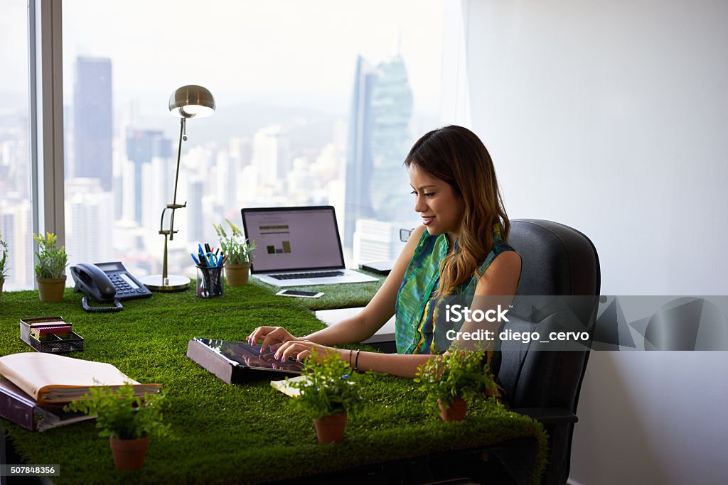Environmentalist Woman Types Email With Tablet On Office Desk Concept of ecology and environment: Young business woman working in modern office with table covered of grass and plants. She types on tablet pc Environmental Conservation Stock Photo