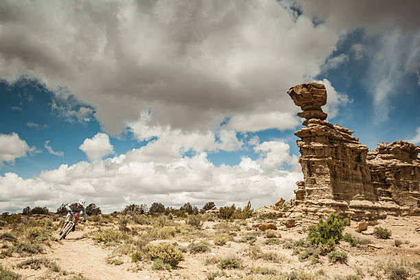 a man pedals his mountain bike while cycling in solitude.  the sunshine creates a lens flare in the desert landscape.  horizontal wide angle composition with copy space.  sanida mountains, albuquerque, new mexico.   pedals past rock formations and is silhoutted by a cloud filled blue sky.  horizontal composition taken in gallup, new mexico.