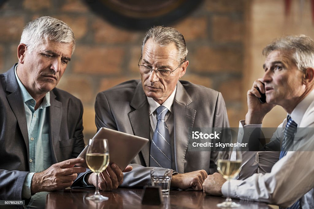 Businessmen in a bar after work. Three businessmen in a bar after work. Two of them are using digital tablet, while the third one is on the phone.   Active Seniors Stock Photo