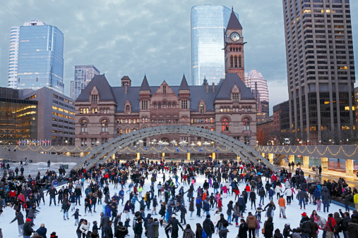 Holiday crowd ice-skating in Toronto's Nathan Phillips Square with Toronto City Hall and Courts plus skyscrapers in the background.  The ice skating rink is adorned with Christmas and New Year decorations.  Moody sky.  Horizontal, copy space, small amount of noise evident due to 800ISO.