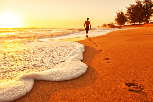 Photo of footprints in a sandy Hawaiian beach at sunset, leading to the silhouette of a surfer walking into the water with his surf board; Kauai, HI.