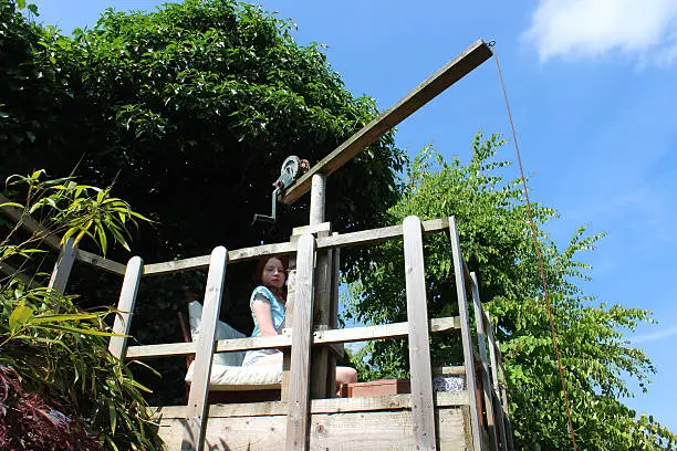 Photo showing a young girl sitting on her treehouse platform, made with fencing posts and decking timber.  A picket style fence provides a safety barrier, while a wooden crane toy using a yacht ratchet device is used to safely raise food and toys up and down from the elevated platform.