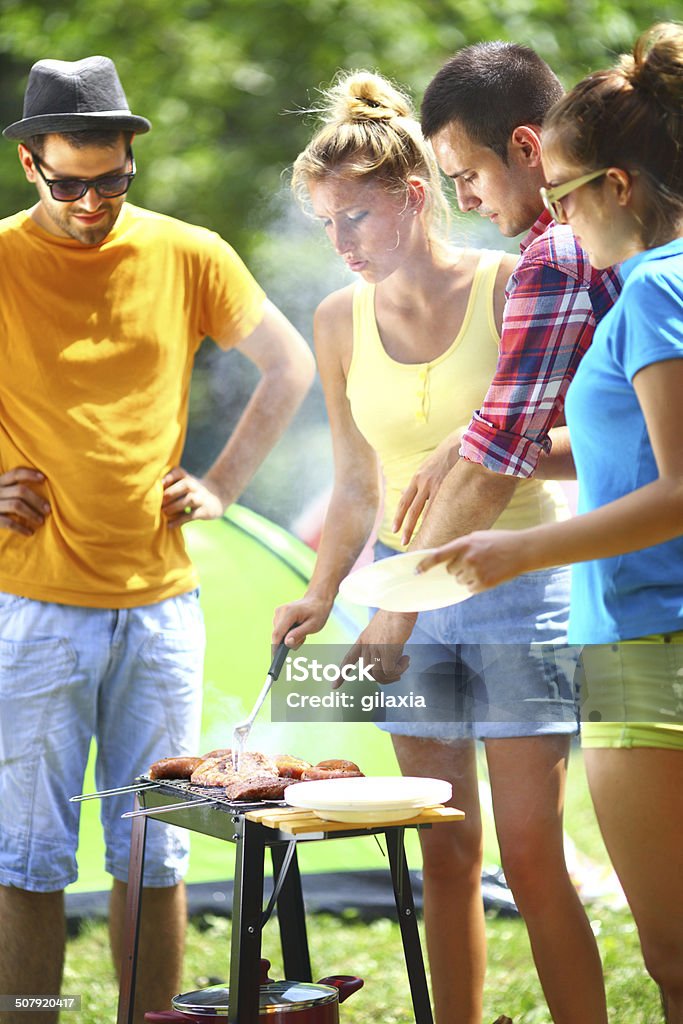 Barbecue party. Group of young adults having fun while grilling food.They are at remote location outside the city.It's sunny summer day.There are two guys and one girl being carried on shoulders..One guy is a chef while the rest are standing by,hungry. All of them wearing summer casual clothes. Barbecue Grill Stock Photo