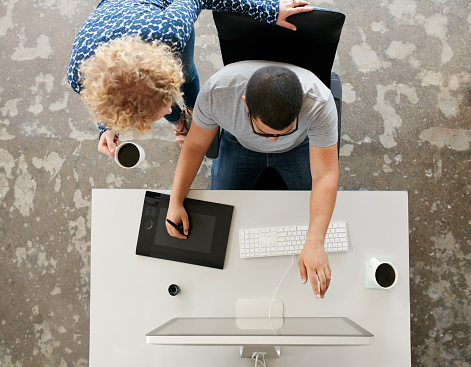 Top view of young graphic designers working in office. Using digitized graphic tablet, digitized pen and desktop computer. Man showing his work on monitor to woman standing by with a cup of coffee.