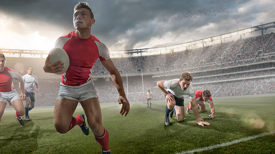A close up image of a heroic looking professional rugby player holding ball and running past opposition players and teammates during a game of rugby. The action occurs during a rugby match in a generic outdoor floodlit stadium full of spectators under a dramatic dark evening sky. 
