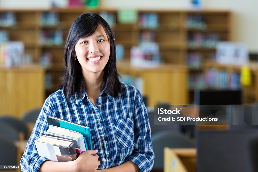 Asian female teacher holding books standing in high school library Pretty Vietnamese teacher or college student is standing in school library holding a stack of textbooks. She has black hair and is looking at the camera and smiling. She is wearing a blue plaid shirt. Desktop computers and bookshelves are in the background. Teacher Stock Photo