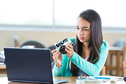 Pretty Hispanic high school student in STEM school works on robot in after school engineering club. She is looking at instructions on her laptop as she assembles the robot. She is concentrating as she looks at the laptop. She is wearing trendy layered clothing and has long brown hair.