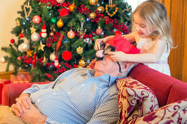 Christmas Fun A cheeky little girl puts funny glasses on her grandfather while he sleeps on the sofa. sleeping grandpa stock pictures, royalty-free photos & images