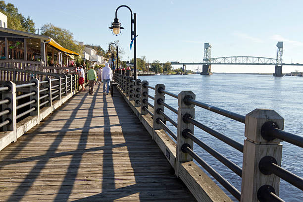 People Stroll Along Water Street Boardwalk, Wilmington, NC Wilmington, North Carolina, USA-November 13, 2015: People stroll along the downtown boardwalk on Water Street alongside tour boats on the water and benches cape fear stock pictures, royalty-free photos & images