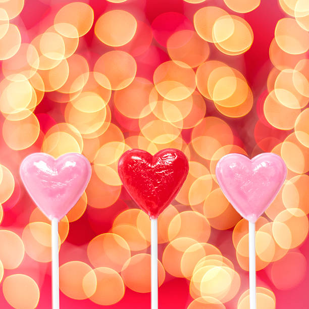 Row of heart shaped lollipops and bokeh on red background A row of three heart shaped lollipops, two pink and one red with amber and red bokeh on red background for Valentine's day. menage trois stock pictures, royalty-free photos & images