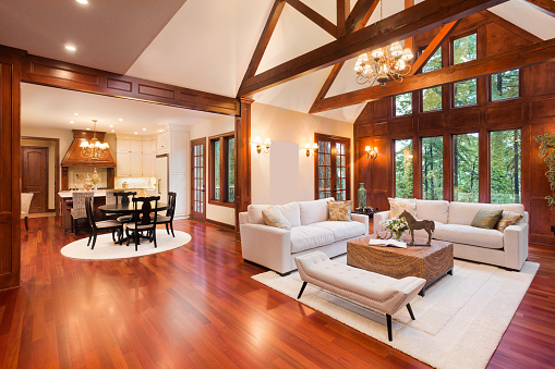 living room interior in luxury home with tall vaulted ceilings, beautiful dark hardwood floors, trim, and trusses, and view of kitchen. Lush green trees make up exterior view.