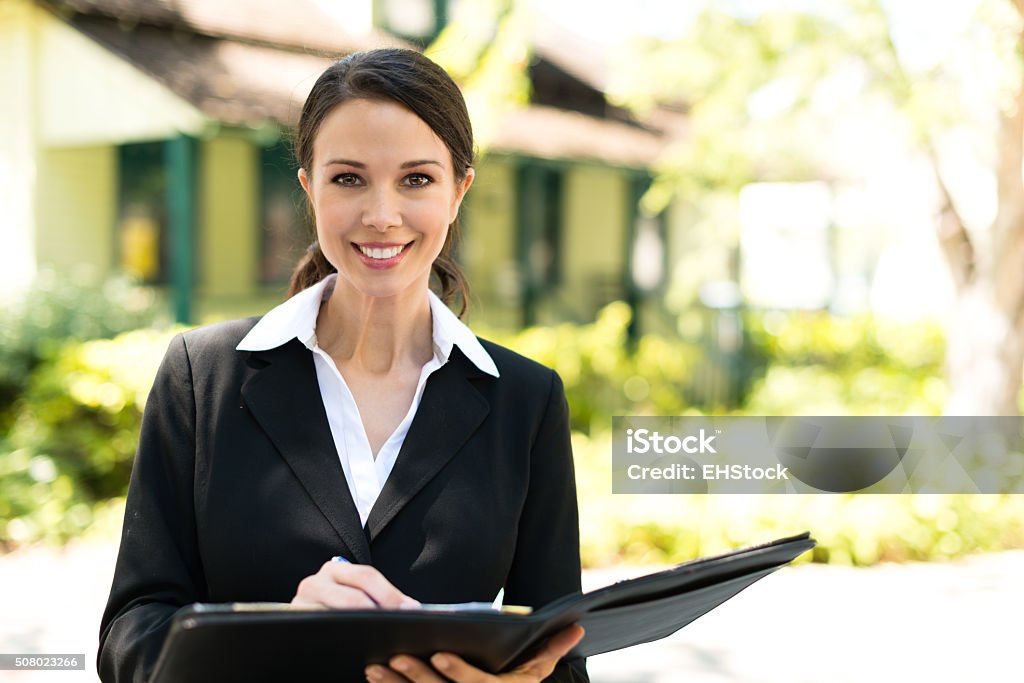 Smiling Real Estate Agent Twenty-something brunette female real estate agent hugging day planner in front of bungalow House Stock Photo