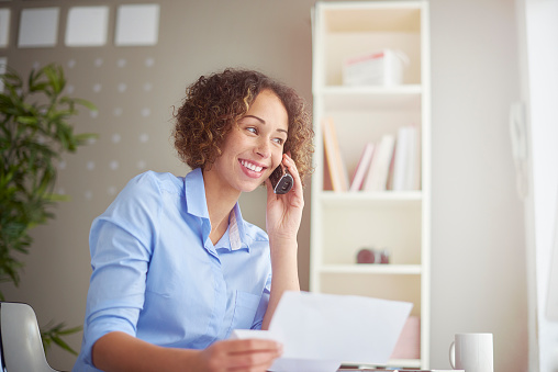a business owner sits at her desk holding a bill and speaking on the phone to her bank or service provider. her expression is happy and positive.