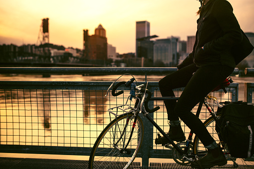A young woman commuting in an urban city environment on her street bicycle takes a break to watch the sunset behind city buildings.  This is the Eastbank Esplanade in Portland, Oregon, that follows along the Willamette river.  