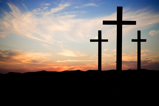 Easter.  The crucifixion.  Three wooden crosses in silhouette stand silently on a hill at sunset.  The dramatic sky is beautiful in its blue and orange colors.  Christianity, religious themes. Copyspace.